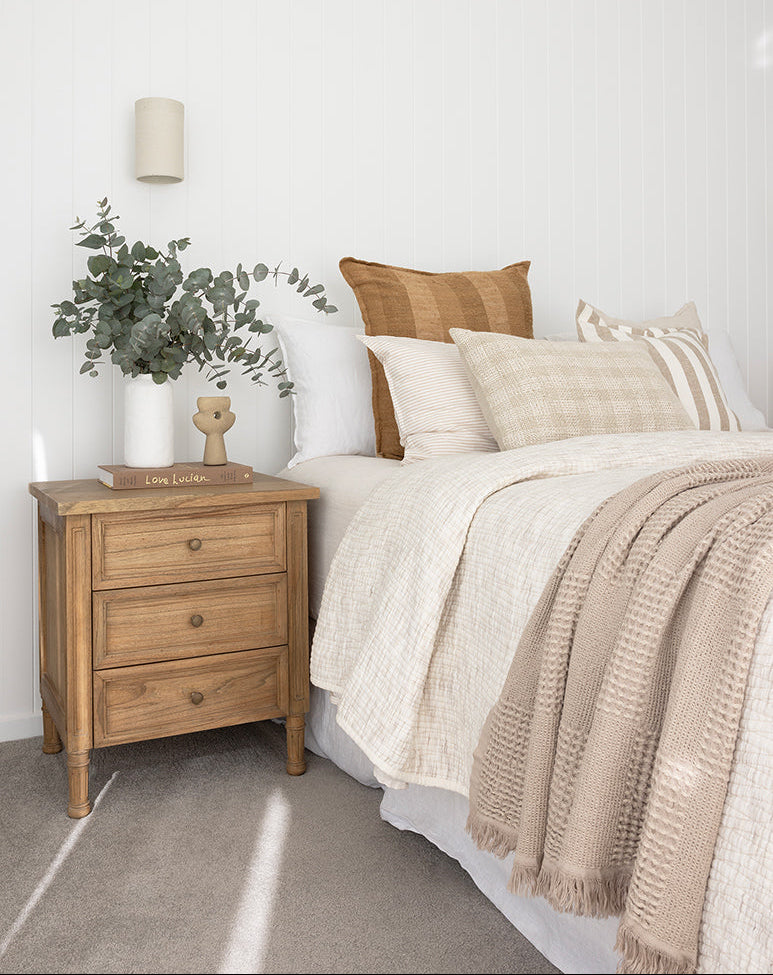 Whitsunday 3 drawer bedside table in weathered oak finish, styled beside a bed with neutral linen bedding and greenery