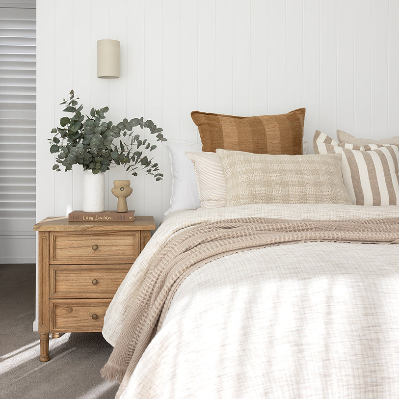 Whitsunday 3 drawer bedside table in weathered oak finish, styled with white bedding, a cushion and greenery