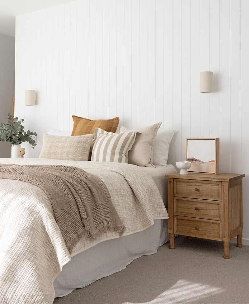 Whitsunday 3 drawer bedside table in weathered oak finish, beside a bed with a framed picture on top