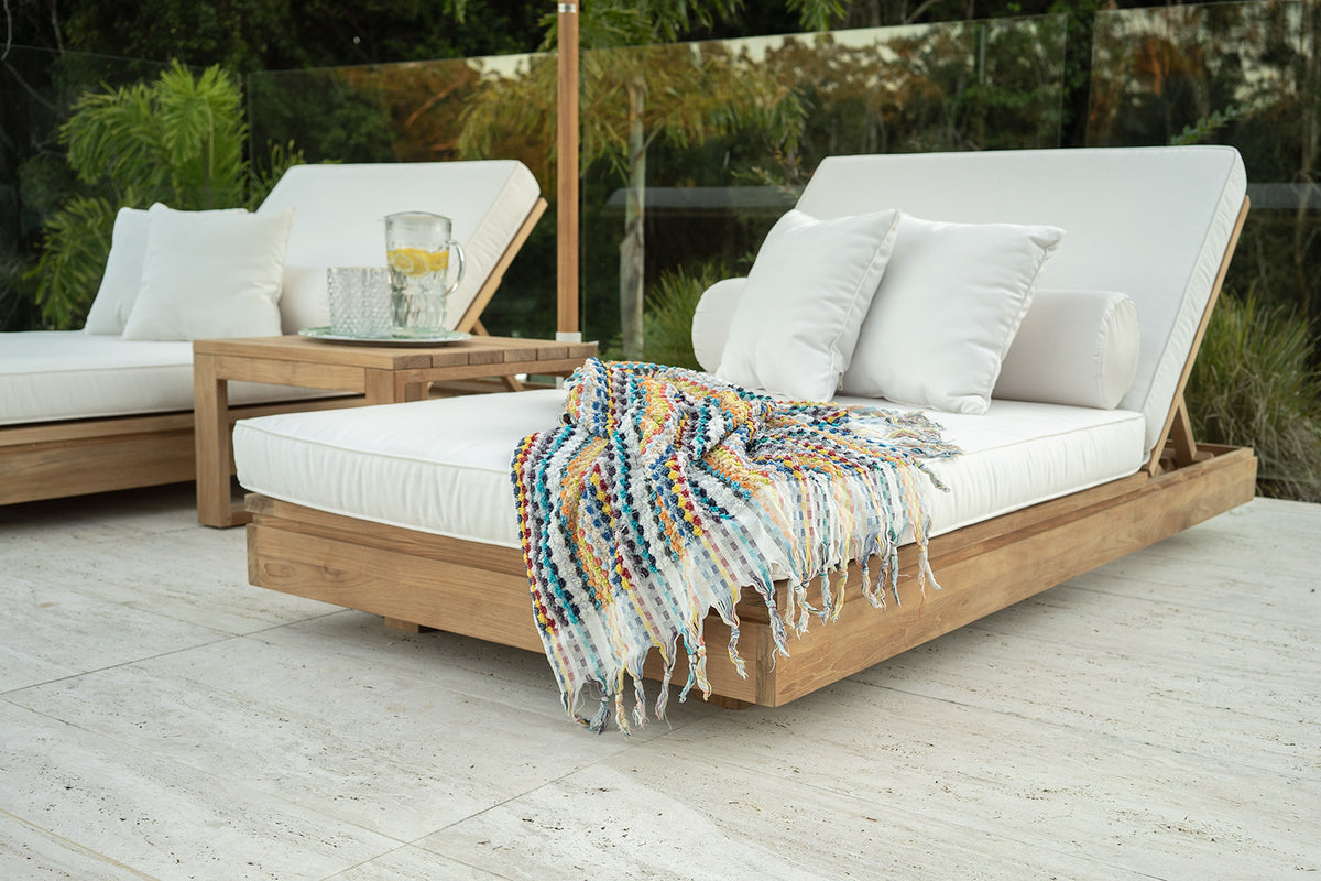 Harbour Island teak sun loungers with light cushions and a patterned throw on a terrace beside greenery