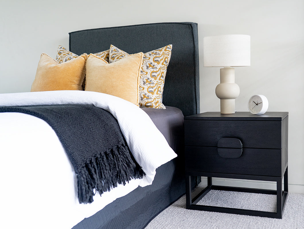 Benji black teak bedside table styled beside an upholstered bed with a white table lamp