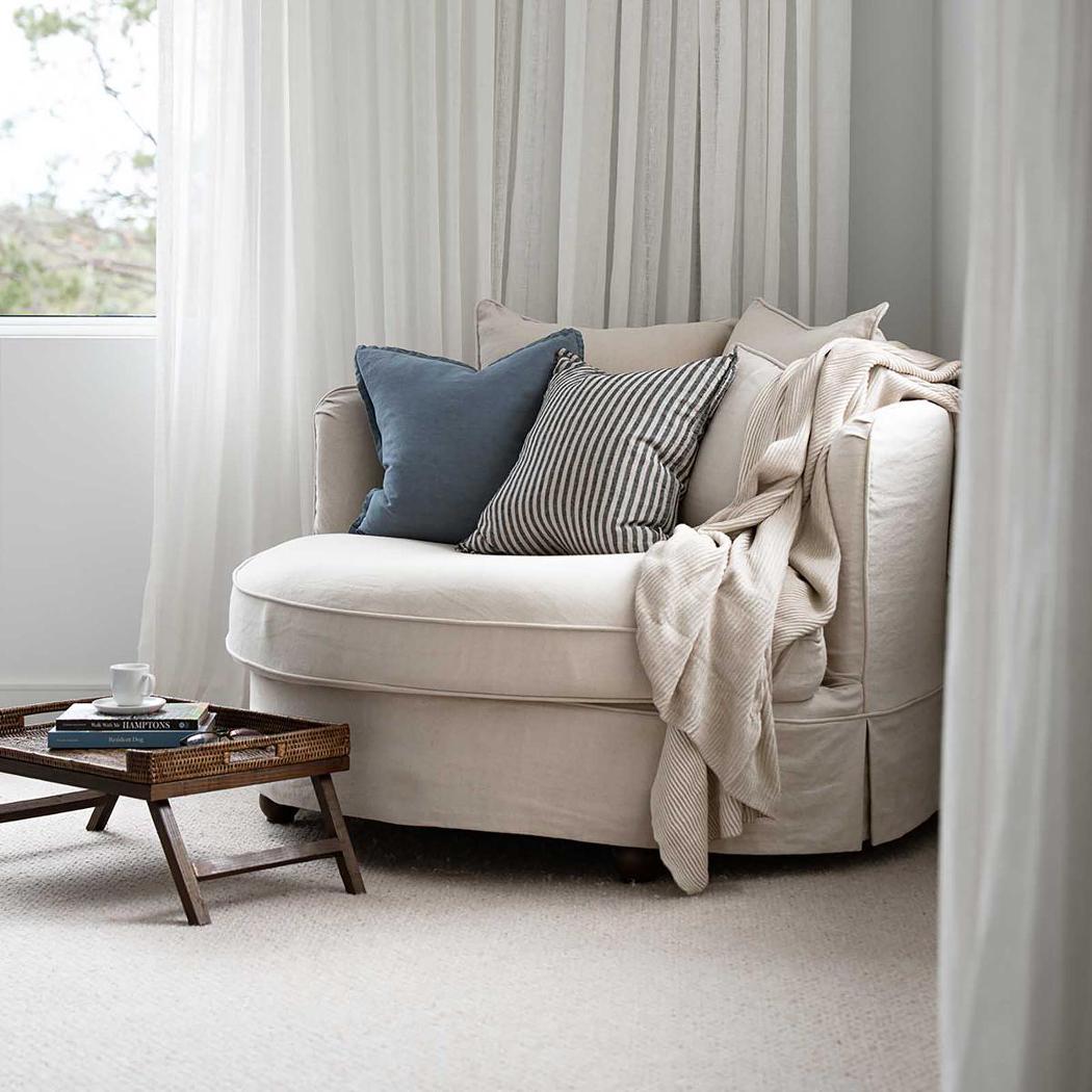 Noosa beige snuggle chair styled beside a window with curtains, cushions and a throw